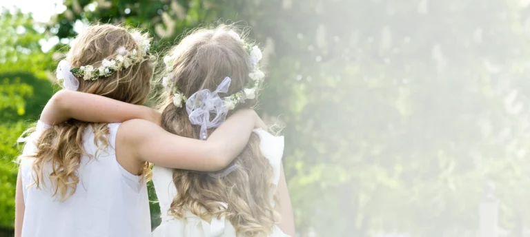 Vestidos de niña - Dos niñas, de espaldas, con sendos brazos pasados por sus hombros, con vestidos blancos elegantes y tiaras de flores